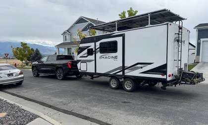 Black GMC Sierra pickup towing an Imagine XLS travel trailer on a suburban street near houses.