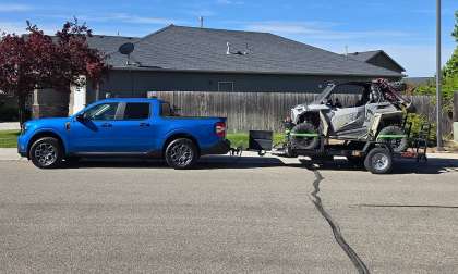 Blue Ford Maverick towing a side-by-side off-road vehicle on a trailer in a residential street.