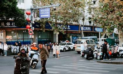 Busy city crosswalk in China with pedestrians, scooters, parked mopeds, and storefronts.