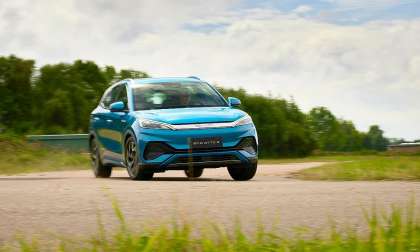 Blue BYD Atto 3 electric SUV driving on a rural road with trees and grass in the background.