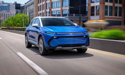 Blue Chevrolet Equinox EV with a white roof driving on an urban road past brick office buildings.