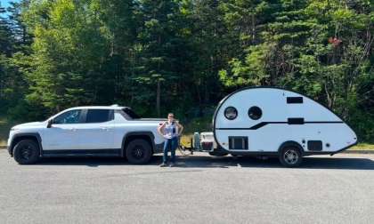 White Chevrolet Silverado EV towing a small white teardrop camper trailer, parked at a forested campsite