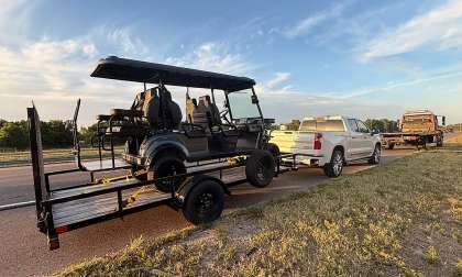 White Chevrolet Silverado towing a golf cart trailer on the roadside with a tow truck parked ahead.