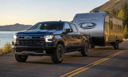 Black Chevrolet Silverado pickup towing a Grey Wolf travel trailer on a road beside a lake and mountains