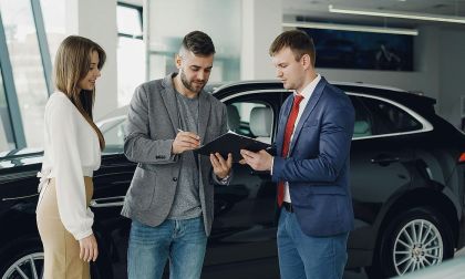 Couple signing car purchase paperwork with male sales agent in blue suit at auto dealership showroom