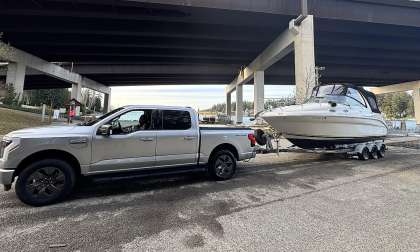Ford F-150 Lightning towing a large cabin boat on a trailer under a bridge near the water.
