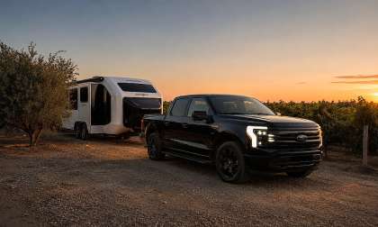 Black Ford F-150 Lightning towing a white Pebble Flow travel trailer through a vineyard at sunset.