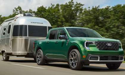 Green Ford Maverick pickup truck towing a silver Airstream travel trailer on a tree-lined highway