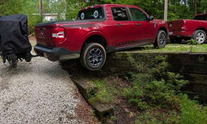 Red Ford Maverick Hybrid XLT towing a covered motorcycle trailer, rear view driving over a low stone retaining wall