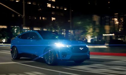 Blue Ford Mustang Mach-E electric SUV driving at night on a city street with headlights on.