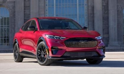 Red Ford Mustang Mach-E electric SUV parked in front of a modern building, front three-quarter view.