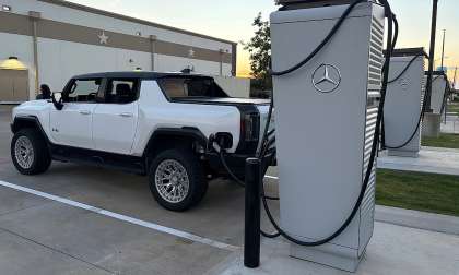 White GMC Hummer EV pickup charging at a Mercedes-Benz charging station at sunset.