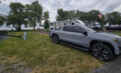Silver GMC Sierra EV Denali pickup truck parked at a lakeside campground, with a Bullet travel trailer and lake visible in the background