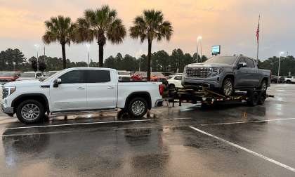 White GMC Sierra pickup towing another GMC Sierra on a trailer in a wet dealership parking lot.