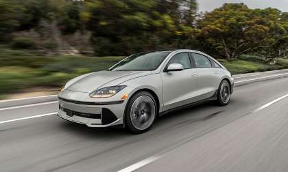 White Hyundai Ioniq 6 driving on a tree-lined road with motion blur and a cloudy sky.