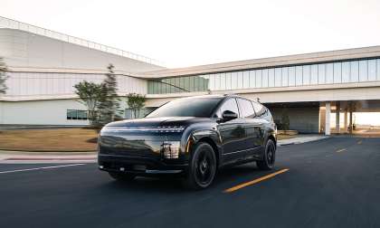 Black Hyundai Ioniq 9 driving past a modern building campus, shown from the front three-quarter angle.