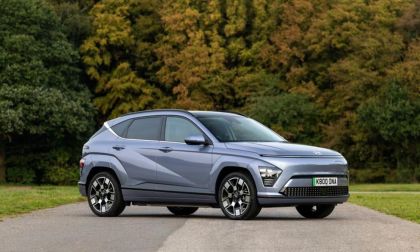 Grey Hyundai Kona Electric parked on a rural road at sunset, shown from a front three-quarter angle with a colorful sky.