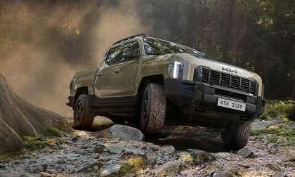 Kia Tasman pickup driving over rocky terrain on a forest trail, kicking up dust during off-road driving.
