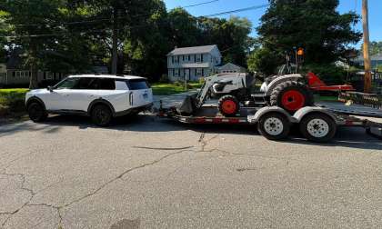 White Kia Telluride SUV towing a flatbed trailer loaded with a compact tractor on a suburban street