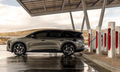 Lucid Gravity electric SUV charging at a Tesla Supercharger station, side profile with multiple charging stalls in a desert landscape.