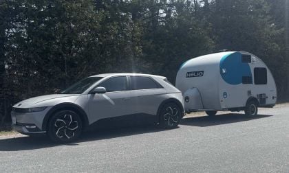 A 2025 Hyundai Ioniq 5 towing a compact, modern camper trailer on a quiet road, framed by tall green trees.