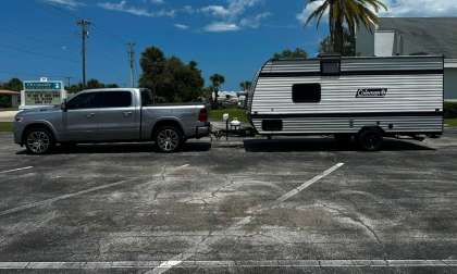 Silver Ram 1500 pickup towing a small Coleman travel trailer in a parking lot with palm trees