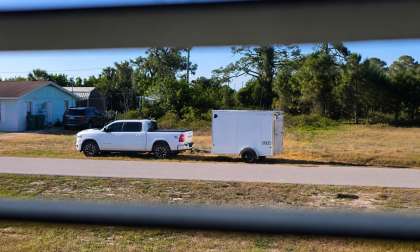 White Ram 1500 pickup towing a small enclosed cargo trailer, parked on a roadside near trees and a house