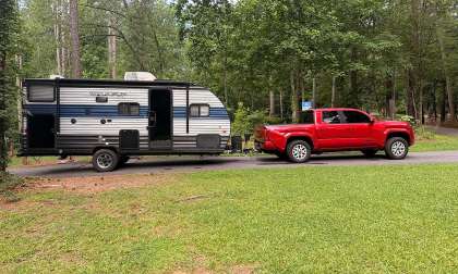 Red Toyota Tacoma towing a Wolf Pup travel trailer on a wooded driveway near a campsite.