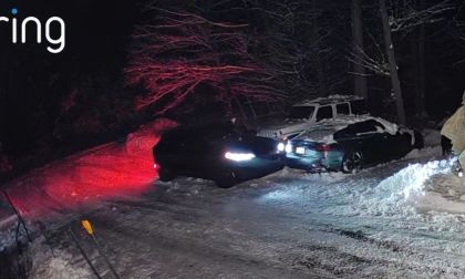 Ring camera footage at night showing a dark Ford Mustang Mach-E with bright headlights and red taillights, and a dark sedan, stuck or maneuvering in a snow-covered driveway.