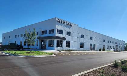 Rivian facility exterior with a large Rivian sign on the building, parked vehicles, and an empty parking lot under a clear blue sky.
