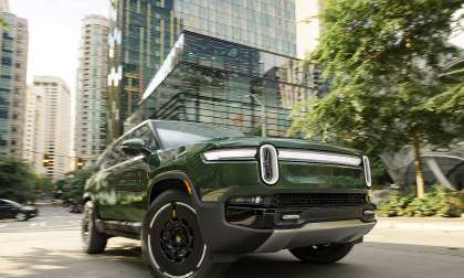 Green Rivian R1S driving through a downtown city street, shown from a low front three-quarter angle.