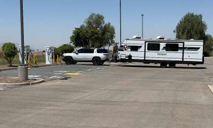 White Rivian R1S and Passport travel trailer shown in side profile at an EV charging station.