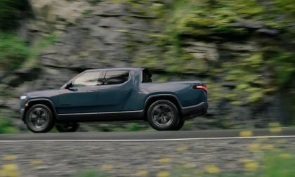 Side view of a Rivian R1T electric truck driving on a road with a rocky, green hillside background, motion blur showing speed.