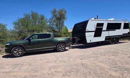 A green Rivian R1T electric pickup towing a white and black Brinkley travel trailer on a gravel lot.