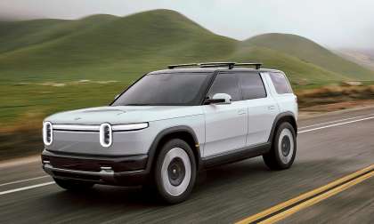 Rivian R2 electric SUV driving on a two-lane road with rolling green hills in the background.