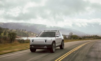 White Rivian R2 electric SUV driving head-on on a two-lane highway under dramatic overcast skies with green hills in background