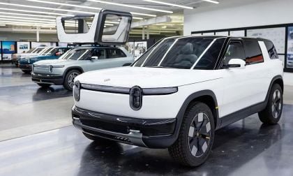 White Rivian R2 compact electric SUV concept on display in Rivian design studio alongside teal R2 and R1S prototypes with design sketches on wall in background