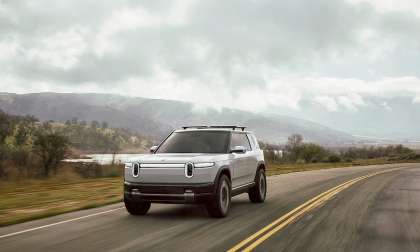 White Rivian R2 driving on a two-lane road with cloudy hills in the background.