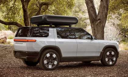 White Rivian R2 parked under trees with a rooftop cargo box in a side-profile view.