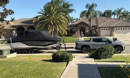 Silver GMC Sierra EV towing a covered boat past large homes and palm trees in a residential neighborhood.