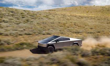Tesla Cybertruck driving fast on a dusty off-road trail through rolling hills under a cloudy sky.