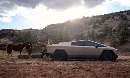 Tesla Cybertruck parked at a ranch beside horses and hay bales, shown from the side profile with hills in the background.