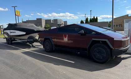 Tesla Cybertruck towing a MasterCraft boat while parked beside Tesla Supercharger stalls in a sunny parking lot.