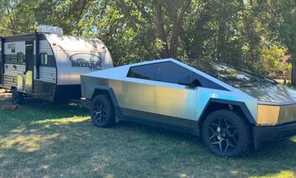 Tesla Cybertruck towing a Patriot Edition travel trailer, parked on grass under shade trees with a dog resting nearby