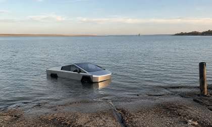 Tesla Cybertruck driving through shallow water at a boat ramp near a lakeshore.