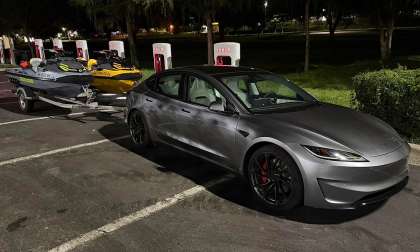 Tesla Model 3 towing two Sea-Doo personal watercraft on a trailer at a Tesla Supercharger station at night.