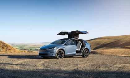Blue Tesla Model X electric SUV with falcon-wing doors open, parked on a scenic overlook in a rural landscape.
