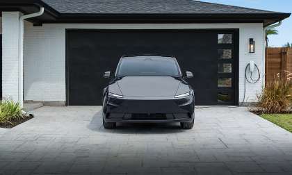 Front view of a dark Tesla Model Y parked in a modern driveway in front of a black garage door with a wall charger mounted nearby.