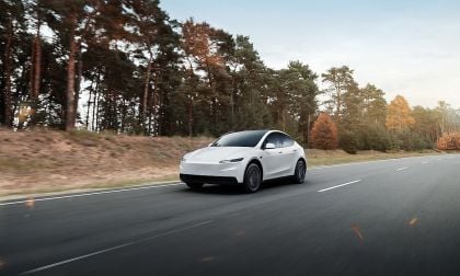 Refreshed Tesla Model Y in white driving head-on on European highway with autumn pine forest in background