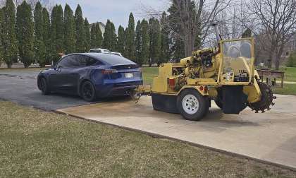 Blue Tesla Model Y towing a yellow construction trencher on a small trailer in a parking lot.